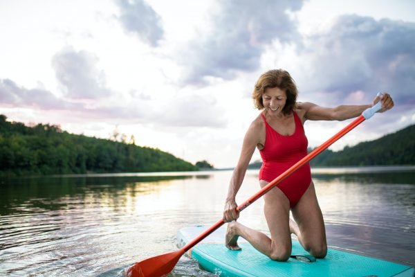 Senior woman paddleboarding on lake in summer. Copy space.
