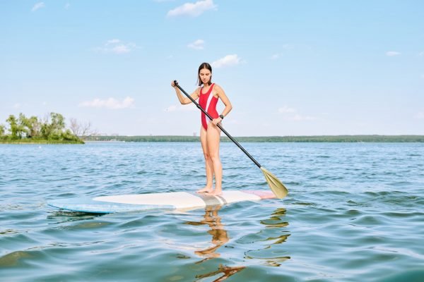 Young pretty brunette sportswoman in red swimsuit floating on surfboard